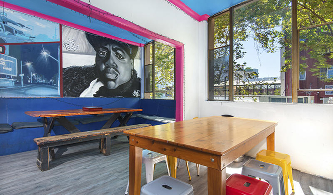 Common area with wooden tables, bright stools, and murals on the wall at Mad Monkey East Side (formerly Hump Backpackers) in Sydney, Australia